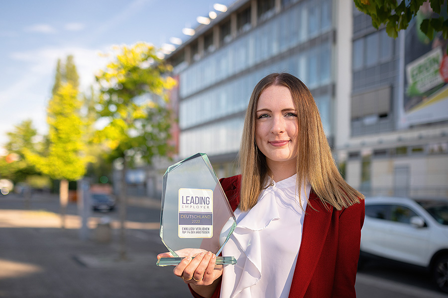 Das Bild zeigt das Portrait einer jungen Frau in weißer Bluse und rotem Blazer, die in die Kamera lächelt. In der Hand hält sie eine gläserne Trophäe mit der Aufschrift "Leading Employer Deutschland 2020".