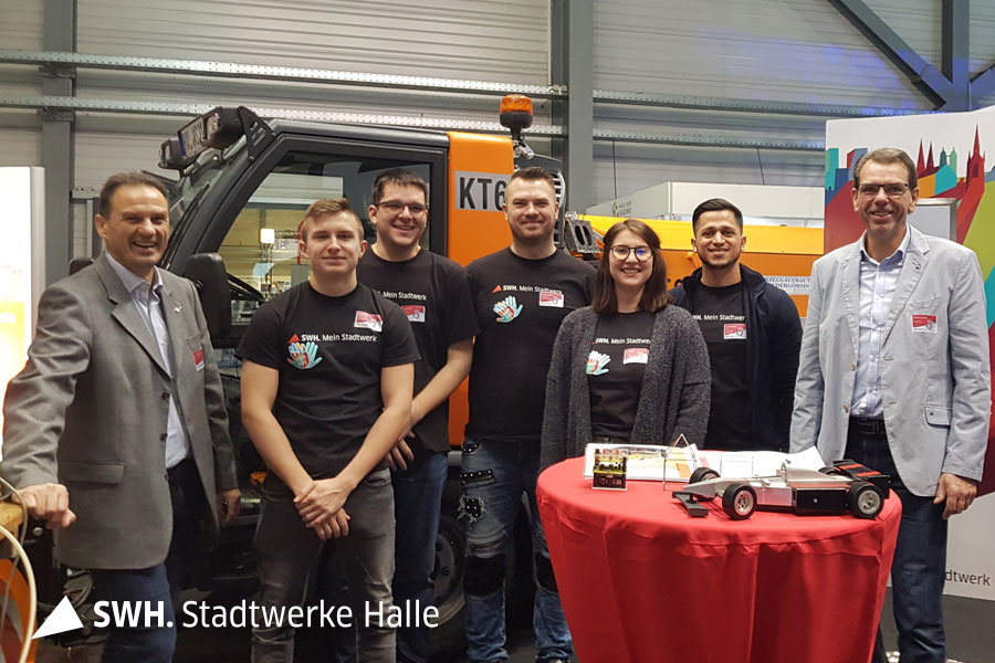 Sieben Personen stehen in einer Halle vor einen kleinen Kehrmaschine. Vor ihnen steht ein kleiner Tisch mit einer roten Husse.