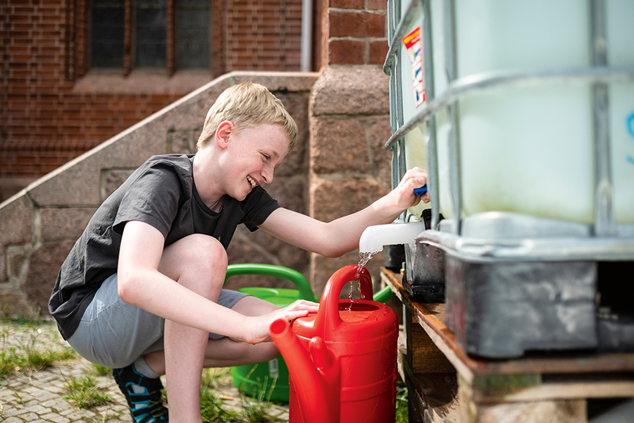 Auf der rechten Bildhälfte befindet sich ein großer Wasserkanister, auf der linken Seite kniet ein blonder Junge, der Wasser aus dem Kanister in eine rote Gießkanne füllt.