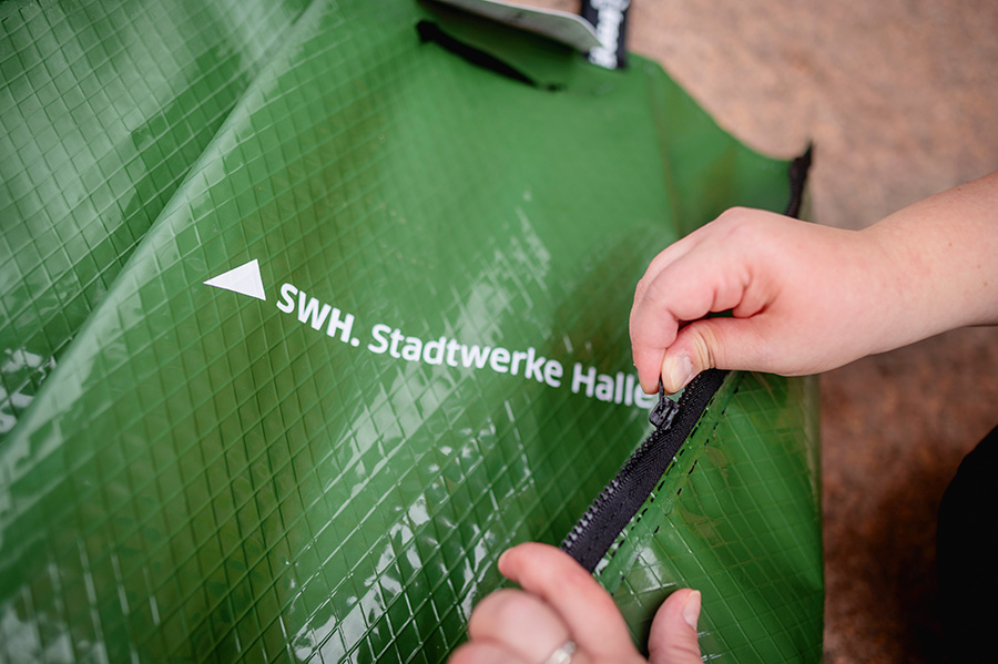 Das Bild zeigt einen Ausschnitt eines grünen Bewässerungssacks mit der weißen Aufschrift "SWH.Stadtwerke Halle". Der Reißverschluss des Sacks wird von zwei Händen geschlossen.