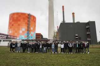 46 Azubis stehen mit Maske nebeneinander. Sie stehen auf einer Wiese. Im Hintergrund ist der Energiepark in der Dieselstraße zu sehen.